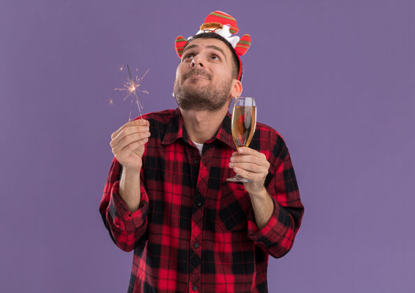 玻璃令人印象深刻的年轻白种人戴着圣诞老人的头带 拿着节日火花和一杯香槟 看着紫色背景上的孤立举行紫色向上