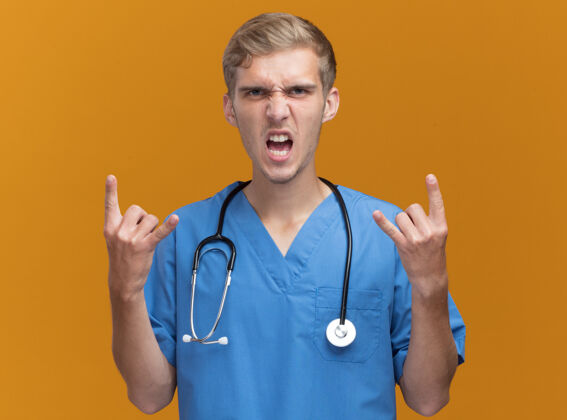 人愤怒的年轻男医生穿着医生制服 带听诊器 在橙色的墙上显示山羊的手势表演表情衣服
