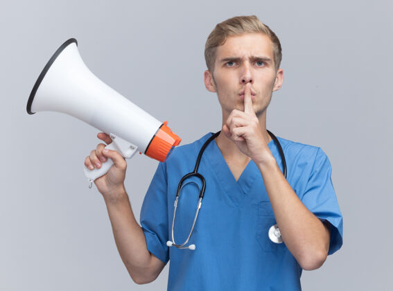 听诊器严格的年轻男性医生穿着医生制服 手持听诊器 在白墙上显示出沉默的手势沉默公民表情