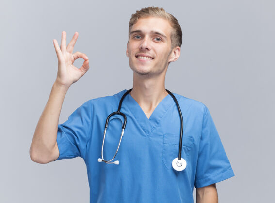 站着微笑的年轻男医生穿着医生制服 听诊器显示出良好的姿态隔离在白墙上衣服手势男性