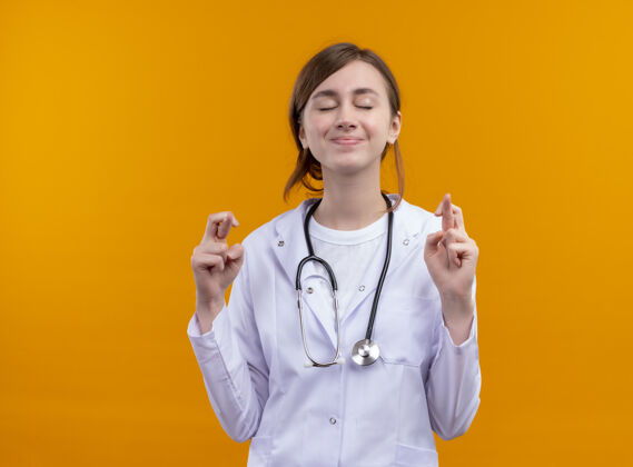 穿梦见年轻的女医生穿着医用长袍和听诊器在孤立的橙色空间里做交叉手指的手势十字架医生长袍