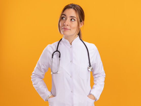 衣服令人印象深刻的一面看年轻女医生穿着医用长袍听诊器把手放在口袋隔离在黄色的墙上印象人人