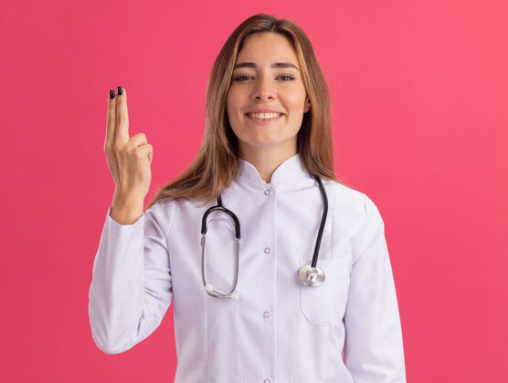 微笑面带微笑的年轻女医生穿着医用长袍 听诊器显示手势 隔离在粉红色的墙上年轻人听诊器人
