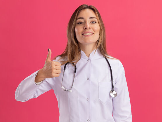拇指面带微笑的年轻女医生穿着医用长袍 听诊器在粉红色的墙上孤立地竖起大拇指年轻人脸听诊器