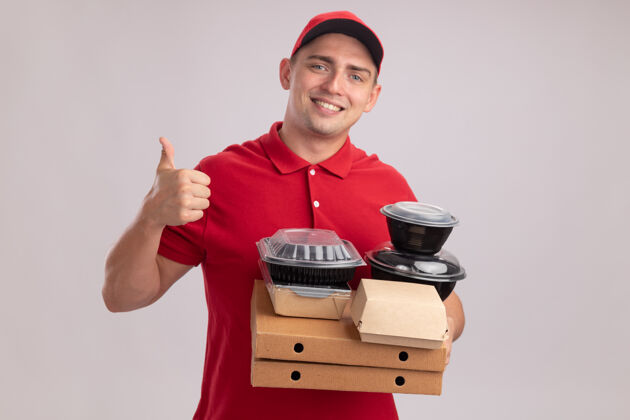拇指微笑的年轻送货员身穿制服 戴着帽子 拿着比萨饼盒上的食品容器 在白色的墙上孤立地竖起大拇指帽子盒子送货
