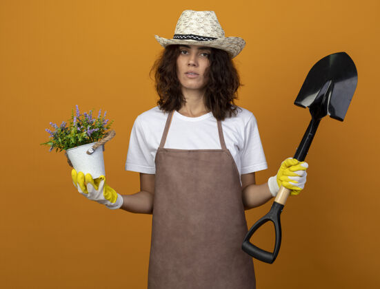 花园穿着制服的年轻女园丁戴着园艺帽和手套 手里拿着花盆里的花和橘子上孤立的铲子花盆女举行