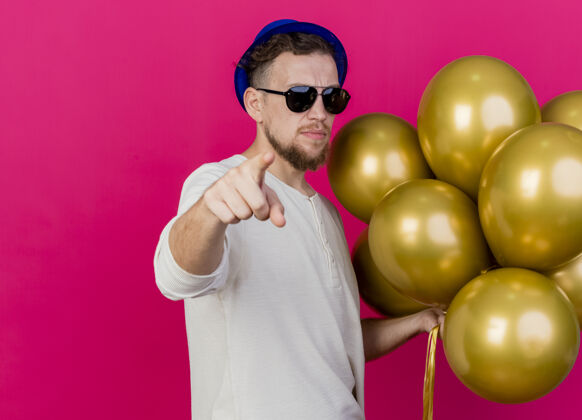 聚会自信的年轻帅哥斯拉夫党家伙戴着党帽和太阳镜举行气球看着和指着前面的粉红色墙上的复制空间孤立太阳镜人公民