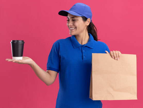 手带着微笑的年轻送货女孩 穿着制服 戴着帽子 手里拿着一个纸制食品包 看着孤立在粉红色墙上的一杯咖啡拿着帽子送货