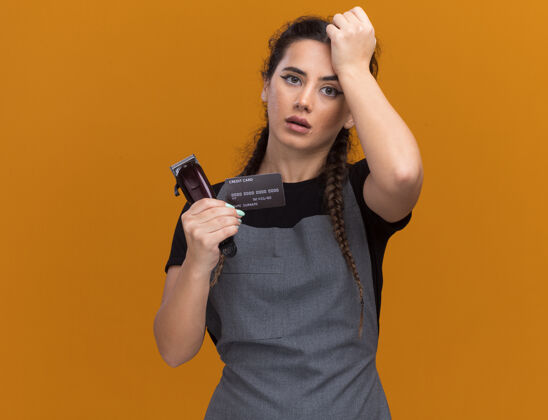 遗憾遗憾的是 年轻的女理发师穿着制服 手里拿着信用卡和理发器 把手放在额上 隔离在橙色的墙上 留着复印空间制服额头信用