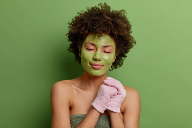 清洁冷静的非裔美国人卷曲浓密的头发拿着沐浴手套包在柔软的毛巾上涂上绿色保湿面膜洗澡大笑女人
