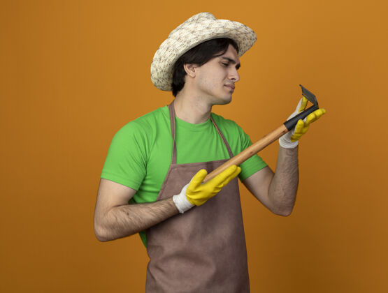 手套年轻的男园丁穿着制服 戴着园艺帽 戴着手套 拿着一把耙子望着孤立的桔子磨损花园年轻