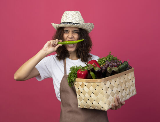 篮子快乐的年轻女园丁穿着制服 戴着园艺帽 手里拿着菜篮子 试着把胡椒粉隔离在粉色上年轻举行园艺