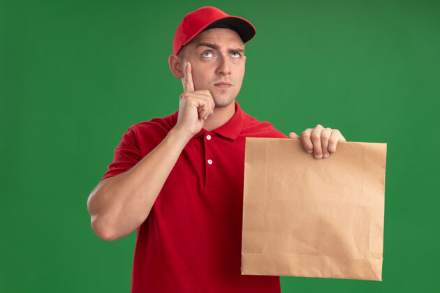 脸颊想想看年轻的送货员穿着制服戴着帽子拿着纸食品包把手指放在脸颊上隔离在绿色的墙上男人帽子思考