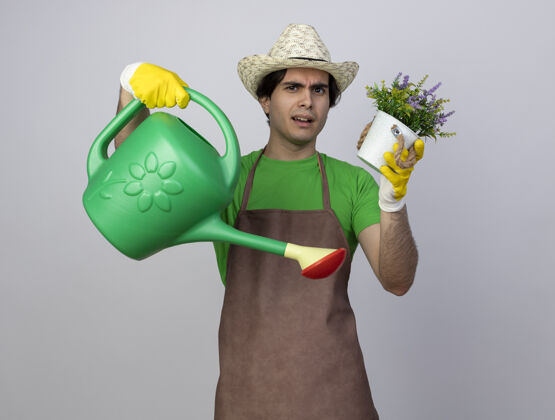 人迷茫的年轻男园丁穿着制服 戴着园艺帽和手套 手里拿着浇花罐 花盆里有花帽子花盆花园