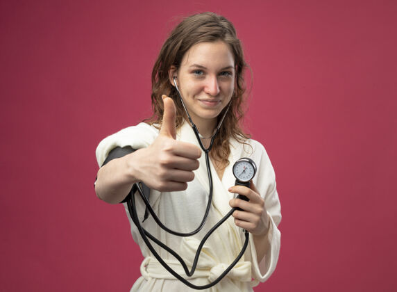 测量穿着白色长袍 用血压计测量自己血压的年轻病女孩高兴地竖起大拇指 孤立地站在粉红色的床上疾病显示穿