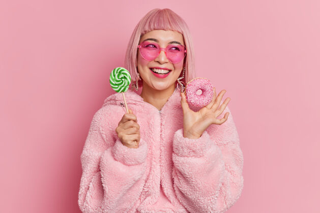 粉色笑容可掬的亚洲少女兴高采烈地把棒棒糖放在棒棒糖和开胃甜甜圈旁边时尚思考请