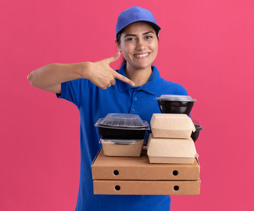 女孩微笑着的年轻送货女孩穿着制服 戴着帽子 指着隔离在粉红色墙上的比萨饼盒上的食品容器制服年轻人披萨