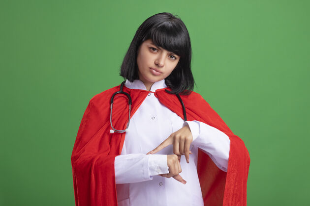 年轻年轻的超级英雄女孩戴着听诊器 穿着医用长袍和斗篷 在绿色的墙上显示着腕表的手势穿手势显示