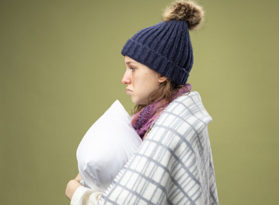 格子悲伤的年轻生病的女孩站在侧视图穿着白色长袍和冬季帽子与围巾包裹在格纹抱枕长袍年轻女人