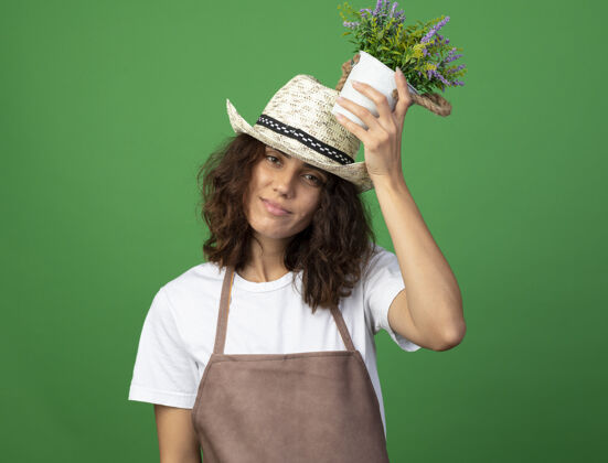 戴高兴的年轻女园丁穿着制服戴着园艺帽把花盆里的花放在头上花盆请制服