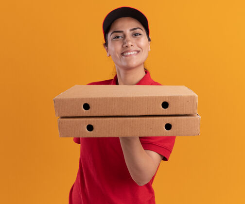 拿着身着制服 头戴鸭舌帽 面带微笑的年轻送货员 把披萨盒孤立在橙色的墙上披萨年轻人穿