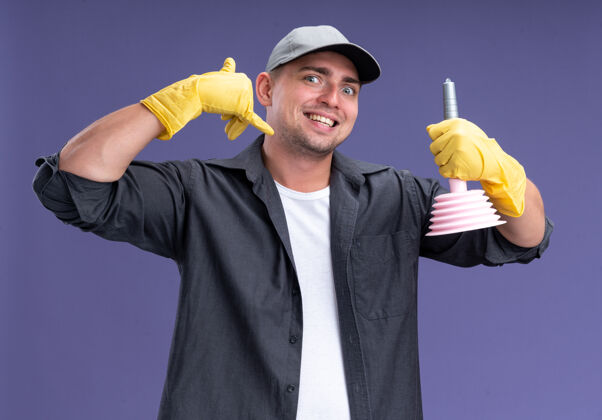 手势兴奋的年轻帅气的清洁工 穿着t恤 戴着帽子 拿着玻璃 在紫色的墙上显示着电话的手势秀抱着帽子