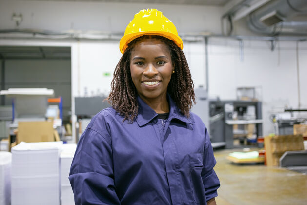 劳动开朗的非洲裔美国女工厂员工戴着安全帽 站在工厂的地板上 面带微笑地看着前面生产工程师微笑