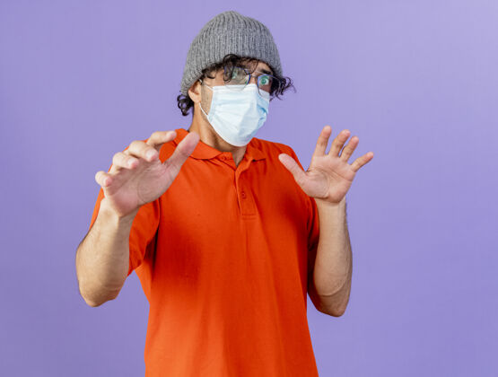 印象印象深刻的年轻病夫戴着眼镜 戴着口罩 戴着冬日帽 望着前方 双手在空气中隔离 在紫色的墙上留着临摹的空间人空气疾病