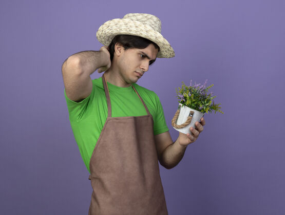 园艺迷茫的年轻男园丁穿着制服戴着园艺帽拿着花盆里的花看着手放在脖子上孤立的紫色花盆帽子紫色