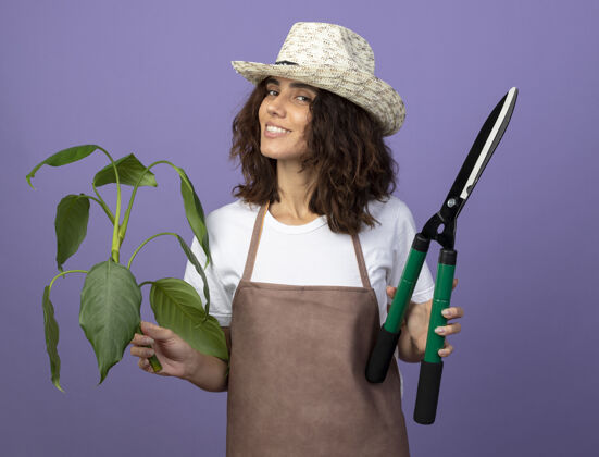 女人微笑着的年轻女园丁穿着制服戴着园艺帽拿着植物拿着剪子年轻紫色成人