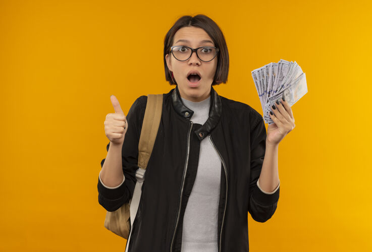 拇指印象深刻的年轻女学生戴着眼镜 背着袋子拿着钱 并显示拇指孤立在橙色橘子背包