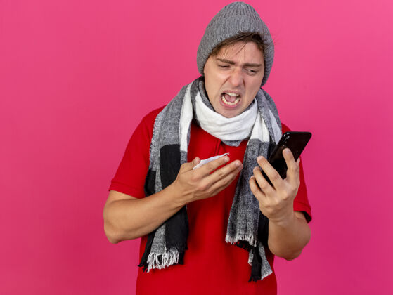 站愤怒的年轻英俊的金发病男子戴着冬天的帽子和围巾拿着手机和餐巾纸看着手机隔离在粉红色的墙上与复制空间表情冬天人