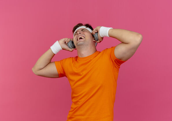 头带快乐的年轻帅气的运动型男人戴着头带 腕带和耳机拿着耳机闭着眼睛孤立在粉色上运动佩戴眼睛