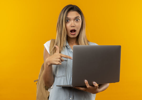 印象印象深刻的年轻漂亮的女学生穿着背袋举行 并指着笔记本电脑孤立的橙色橙色拿着年轻