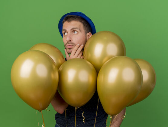 脸体贴的帅哥戴着蓝色的派对帽 手放在下巴上 站着氦气球 看着隔离在绿色墙壁上的一面 还有复制空间气球表情人