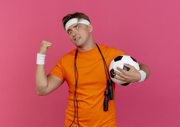 脖子印象深刻的年轻英俊的运动男子戴着头带和手腕与跳绳脖子上举行足球指向后面孤立的粉红色英俊头带绳子
