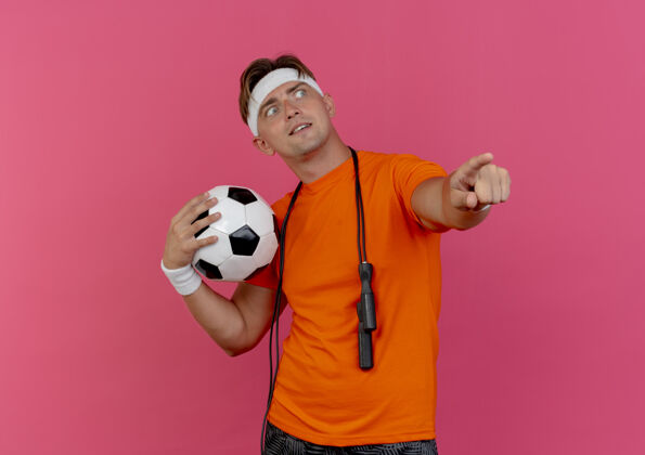 壁板印象深刻的年轻英俊的运动型男子戴着头带和手腕 脖子上系着跳绳 手里拿着足球 指着粉红色的一边看着孤立的男人拿着球