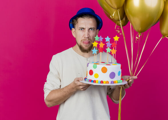 斯拉夫皱眉的年轻帅哥斯拉夫党家伙戴着党帽子拿着气球和生日蛋糕与明星看前面显示舌头孤立在粉红色的墙壁与复制空间人脸市民