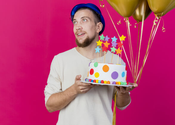 男人令人印象深刻的年轻英俊的斯拉夫党的家伙戴着党的帽子拿着气球和生日蛋糕与明星看一边隔离在粉红色的墙上复制空间印象衣服壁板