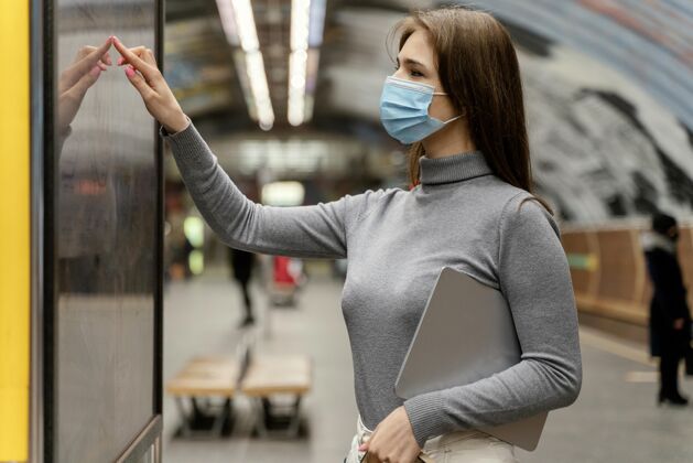女人拿着平板电脑在地铁站等着的年轻女子旅行城市外科口罩
