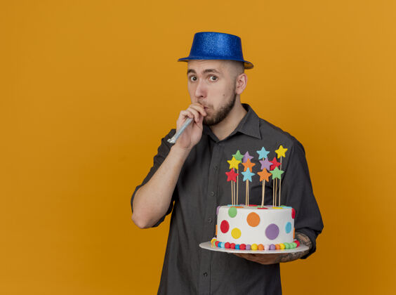 表情令人印象深刻的年轻英俊的斯拉夫党人戴着党帽看着前面举行生日蛋糕吹党鼓风机孤立的橙色墙上与复制空间感觉穿市民