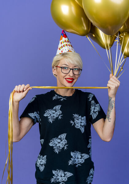 市民微笑着的金发派对女郎戴着眼镜 戴着生日帽 手里拿着气球 孤立地看着前面的紫色墙壁姿势站着穿着