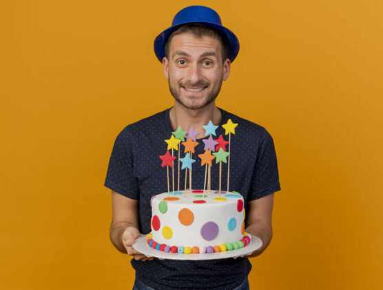 站立微笑的帅哥戴着蓝色的派对帽 拿着生日蛋糕 隔离在橙色的墙上 留着复制空间蛋糕姿势微笑
