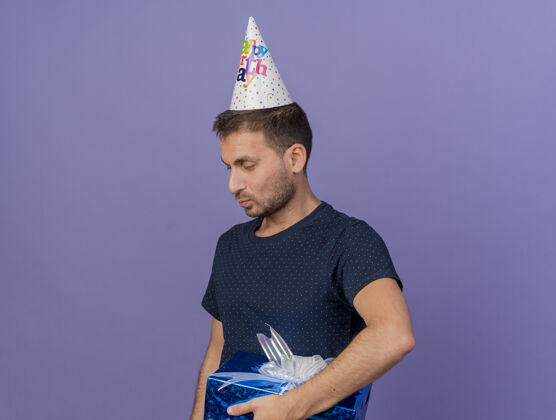 盒子高兴的帅哥戴着生日帽拿着礼盒望着一面隔离在紫色墙上的复制空间感情表情请