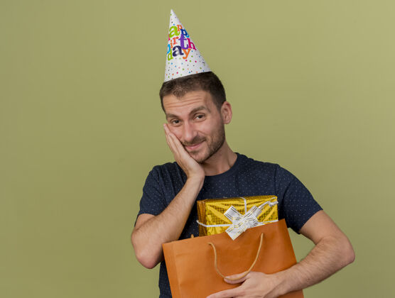 拿着戴着生日帽的帅哥高兴地把手放在脸上 拿着纸购物袋里的礼盒 隔离在橄榄绿的墙上 留着复印空间手衣服礼物