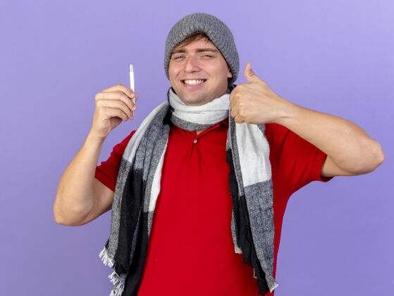 围巾面带微笑的年轻英俊的金发病患戴着冬天的帽子和围巾拿着温度计看着前面伸出大拇指孤立地站在紫色的墙上男人英俊穿着