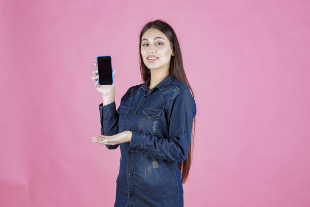 人穿着牛仔衬衫的女孩展示她的智能手机 并竖起大拇指服装喜欢科技