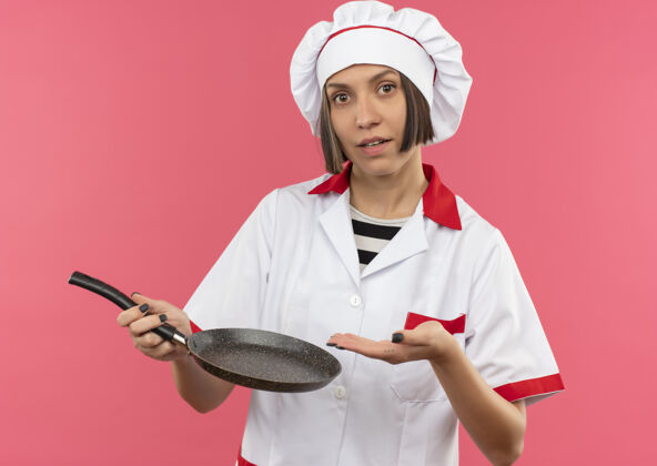 女身着厨师制服的年轻女厨师手拿煎锅 看着隔离在粉色背景上的摄像机手相机厨师