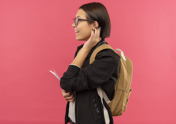 耳朵戴着眼镜 背着书包 站在侧视图里 拿着记事本 抚摸着粉红色的耳朵 快乐的年轻女学生年轻粉红色眼镜