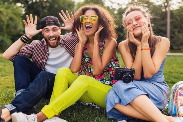 兴奋快乐的年轻朋友们坐在公园里玩着疯狂的笑脸 男人和女人一起玩着 带着相机旅行时尚多种族人